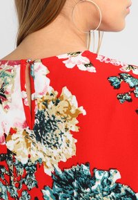 Červená květinová blůza s velkými zelenými a béžovými květy, s otvorem na zádech ve tvaru klíče a hladkým, lehkým materiálem.