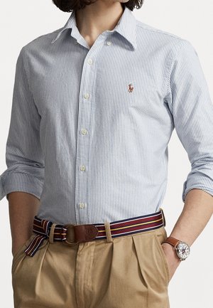 Man wearing light blue striped button-up shirt with rolled sleeves, beige pants, striped belt, and watch with brown leather strap.
