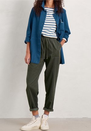 Femme portant une veste bleue par-dessus une chemise rayée, un pantalon vert olive avec cordon, des chaussettes grises et des baskets blanches montantes sur un fond uni.