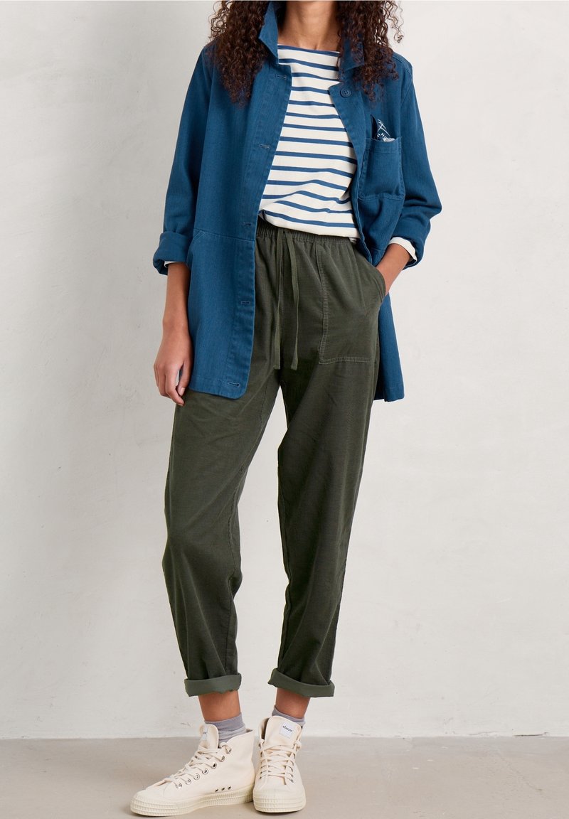 Femme portant une veste bleue par-dessus une chemise rayée, un pantalon vert olive avec cordon, des chaussettes grises et des baskets blanches montantes sur un fond uni.