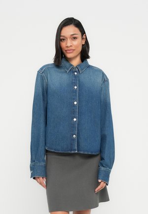 Žena s tamnom kosom do ramena, nosi plavu džins jaknu s gumbima i sivu suknju do koljena, stoji ispred jednobojne pozadine.