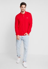 Czerwona polo w długim rękawie z kołnierzykiem, dwoma guzikami i małym zielonym logo. W połączeniu z jasnoniebieskimi dżinsami i białymi trampkami.