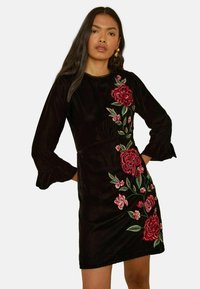 Rochie din catifea neagră cu mâneci lungi, având broderie florală roșie pe o parte și o siluetă conică cu manșete volanate.