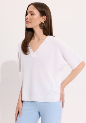 Kobieta z prostymi brązowymi włosami nosząca duże złote kolczyki, biały krótki top z dekoltem w serek i jasnoniebieskie spodnie, pozująca z jedną ręką na biodrze.