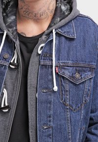 Dunkelblaue Jeansjacke mit einem grauen Kapuzenpullover darunter. Verfügt über silberne Knöpfe, Kordelzüge und Brusttaschen. Strukturierter Stoff.