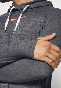 Grau-gefütterter Kapuzenpullover aus strukturierter Baumwolle, mit weißen Kordeln und orangefarbenem Nike-Logo auf der Brust. Mit langen Ärmeln und lässiger Passform.