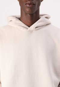 Lysbeige hættetrøje lavet af blødt, tekstureret stof. Har en trekantet halsudskæring og en afslappet pasform med hætte. Simpelt design uden udsmykninger.