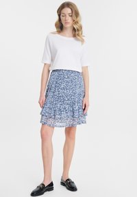 Witte cropped t-shirt gecombineerd met een gelaagde, blauw met bloemenpatroon rok. De rok heeft een doorschijnende overlay en een rucheszoom. Zwarte loafers maken de outfit compleet.