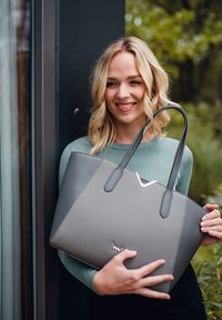 Graue Tragetasche mit doppelt strukturierten Panels und einem silbernen Akzent. Verfügt über robuste schwarze Griffe und ein minimalistisches Logo am Boden.