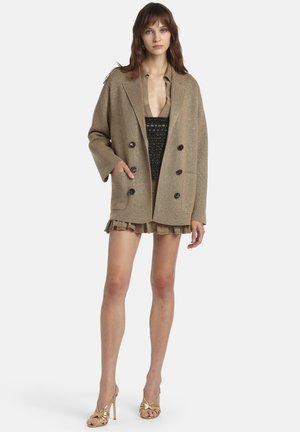 Žena u prevelikom bež dvostruko kopčajućem sakou, bež šortsevima s naborima, crnoj teksturiranoj majici i zlatnim sandalama na visoku petu, stoji nasuprot bijeloj pozadini.