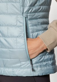 Lyseblå quiltet puffervest med sidelommer med glidelås, med en beige ribbet mansjett på ermene. Glatt tekstur.