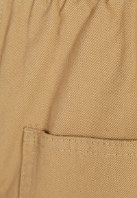 Beige stof met een getextureerd, diagonaal geweven patroon; voorzien van een elastische tailleband en een zijzak met nette stiksels.