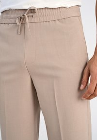 Beżowe spodnie dresowe z kieszeniami bocznymi noszone przez osobę, której widoczna ręka spoczywa przy udzie, na białym tle.