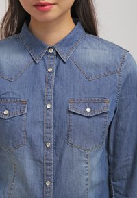 Jeansskjorta i ljusblått med långa ärmar, en spetsig krage, axelsömmar i westernstil och två bröstfickor med tryckknappar.