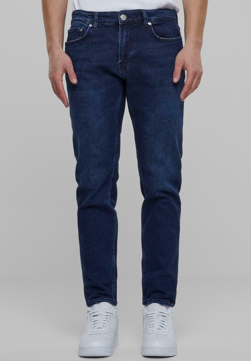 Sötétkék farmernadrág, slim fit szabással, öt zsebes kialakítással és ezüst színű fém kiegészítőkkel. Fehér sportcipővel viselve.