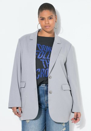Person med kort hår bærer store hoop-øreringe, en grå oversized blazer, en sort t-shirt med blå tekst og revnede blå jeans.
