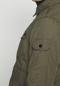 Olivgrön jacka med en strukturerad design, som har en knappkrage, flera påsydda fickor och svarta knappaccenter. Texturerat tyg.