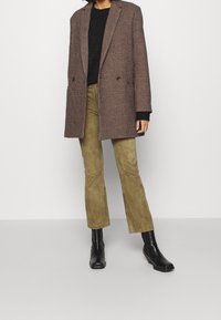 Manteau long marron à carreaux porté sur un pull noir, associé à un pantalon évasé vert olive et des bottines noires. Textures lisses et coupe ajustée.