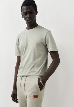 Homme à la peau foncée et aux dreadlocks portant un t-shirt Hugo beige clair et un pantalon de survêtement assorti avec un patch logo rouge, posant devant un fond blanc.