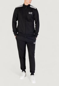 Chándal deportivo negro hecho de tela suave, con una chaqueta con cremallera que presenta detalles a rayas y logo, combinado con pantalones a juego y zapatos blancos.