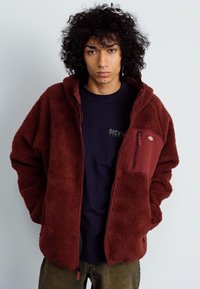 Jachetă fleece bărbie, cu textură moale, cu buzunar fermoar pe piept, închidere frontală cu fermoar și glugă, purtată peste un tricou închis la culoare.
