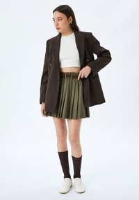 Bruine dubbelgeknoopte blazer over een witte crop top, gecombineerd met een groene plissérok, bruine kniehoge sokken en witte sneakers.