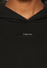 Felpa nera con cappuccio, realizzata in tessuto testurizzato, dotata di tasca a marsupio e un'etichetta rettangolare con "Calvin Klein" in un testo bianco a contrasto.
