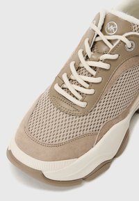 Baskets beige et taupe avec tissu en mesh, superpositions en daim et cuir, lacets blancs, et semelle blanche épaisse, décorées d'un logo MK métallique rond près de la languette.