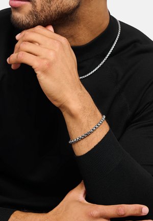 Man wearing black long-sleeve shirt with silver chain necklace and matching silver chain bracelet, hand near chin in a thoughtful pose.