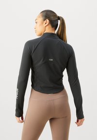 Veste de sport noire à manches longues, au design ajusté, avec un logo dans le dos. Associée à un legging marron taille haute. Texture lisse.