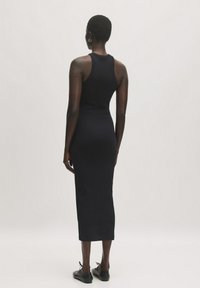 Schwarzes, ärmelloses, geripptes Kleid mit einer taillierten Silhouette, knielang und glatter Textur. Getragen mit schwarzen flachen Schuhen und minimalem Schmuck.