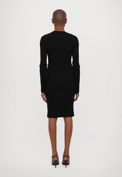 Rochie neagră, ajustată, cu mâneci lungi, cu o siluetă dreaptă, cu tivul la genunchi și textură netedă, asortată cu pantofi cu toc stiletto negri.