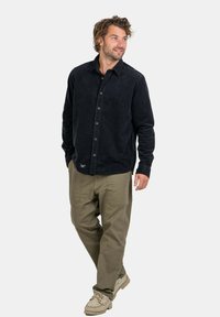 Camicia nera in corduroy con bottoni e colletto, abbinata a pantaloni cargo leggeri color oliva. Dettagli notevoli includono una piccola etichetta con logo e bottoni.