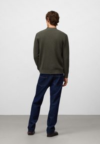 Man som står med ryggen mot kameran, iförd en olivgrön tröja, mörkblå jeans och bruna skor, på en ljusgrå golvyta med vit vägg i bakgrunden.