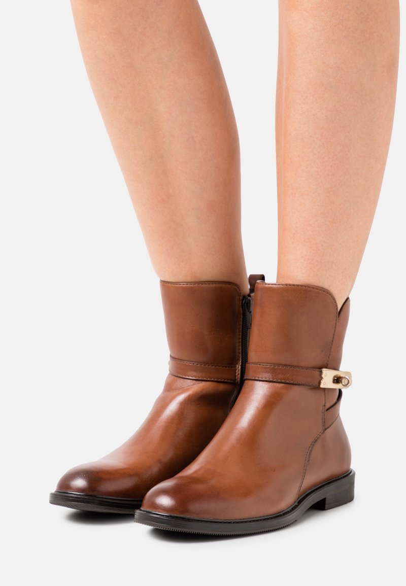 Bottines en cuir marron à fermeture éclair latérale et petite boucle dorée portées sur des jambes nues sur un fond clair.