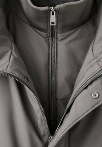 Giacca grigia con zip e colletto a coste. Presenta dettagli in metallo argentato, una texture liscia e cuciture visibili lungo le strutture.