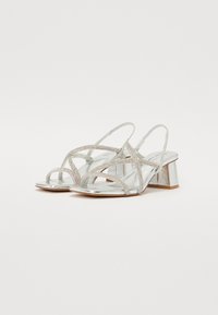 Srebrne metalne slingback pete s križnim trakami in dodatki iz rhinestones; nizka blok peta in gladka, sijoča tekstura.