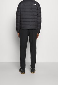 Person wearing black puffer jacket, black pants, and black sneakers standing against a plain light background facing away.