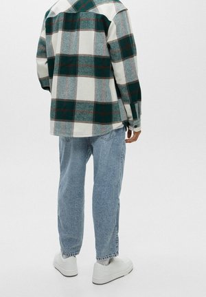 Personne portant une veste à carreaux verte et blanche, un jean bleu clair et des baskets blanches, debout de dos sur un fond uni.