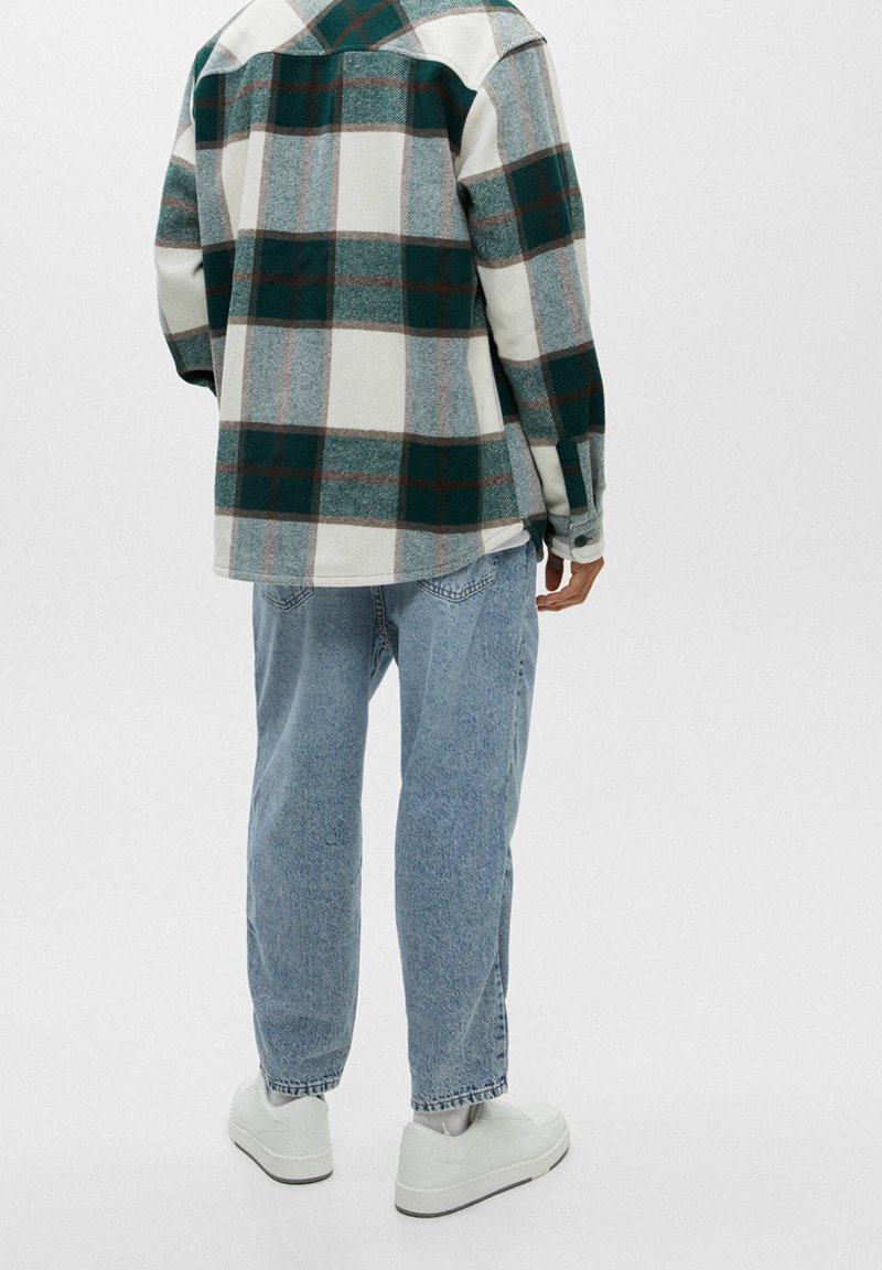 Personne portant une veste à carreaux verte et blanche, un jean bleu clair et des baskets blanches, debout de dos sur un fond uni.