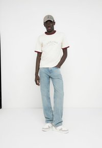 Roomkleurig T-shirt met korte mouwen en bordeauxrode biezen, spijkerbroek en grijs petje. Witte sneakers met lichtgrijze accenten. Staande pose, neutrale achtergrond.