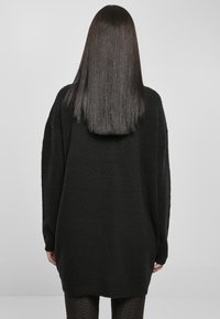 Sweater en tricot noir surdimensionné avec de longues manches, texture douce et ourlet arrondi. Porté sur des leggings sombres à motifs. Vue arrière minimaliste.