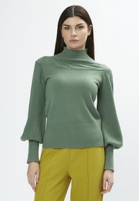 Maglione verde lavorato a maglia con collo alto, spalle con texture e polsini a coste; abbinato a pantaloni gialli sartoriali.