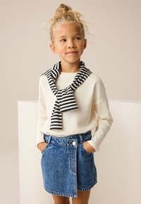 Witte top met lange mouwen en een zwart gestreepte sjaal gestrikt rond de nek, gecombineerd met een blauwe denimrok met een enkele knoop en zijzakken.