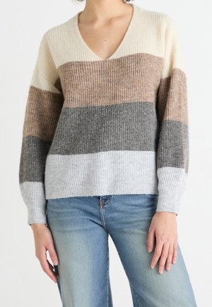 Personne portant un pull à col en V avec de larges rayures horizontales crème, beige, gris et gris clair, associé à un jean bleu clair.
