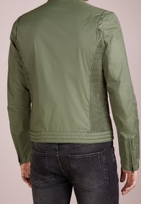 Veste verte légère avec une coupe ajustée. Comprend des panneaux latéraux texturés et des fermetures éclair aux poignets. Finition lisse avec un col montant.
