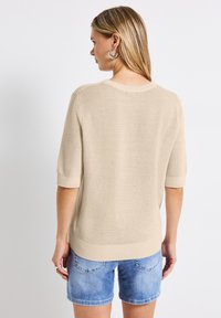 Vaaleanbeige neulepaita, jossa on lyhyet hihat, teksturoitu kuosi ja resorireuna. Yhdistetty vaaleansinisiin farkkushortsseihin.