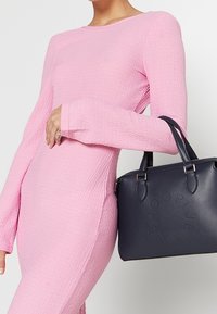 Granatowa torba na ramię ze skóry z wytłaczanym logo, podwójnymi uchwytami i usztywnionym kształtem. Nosi się ją z teksturowaną różową sukienką z długim rękawem.