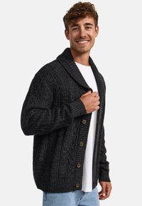 Cárdigan de punto trenzado gris oscuro con cuello de chal y botones marrones, que presenta un acabado de puños y dobladillos acanalados, llevado sobre una camiseta blanca.