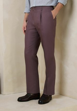 Mand iført brune skræddersyede bukser med læg og sorte læder loafers, står mod en beige væg med den ene hånd i lommen.
